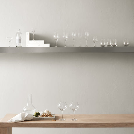 A minimalist dining scene with two Orrefors wine glasses, a decanter, and snacks on a wooden table; an array of glassware and Orrefors: More Champagne (4-Pack) 6 oz glasses line a floating shelf against a neutral wall.