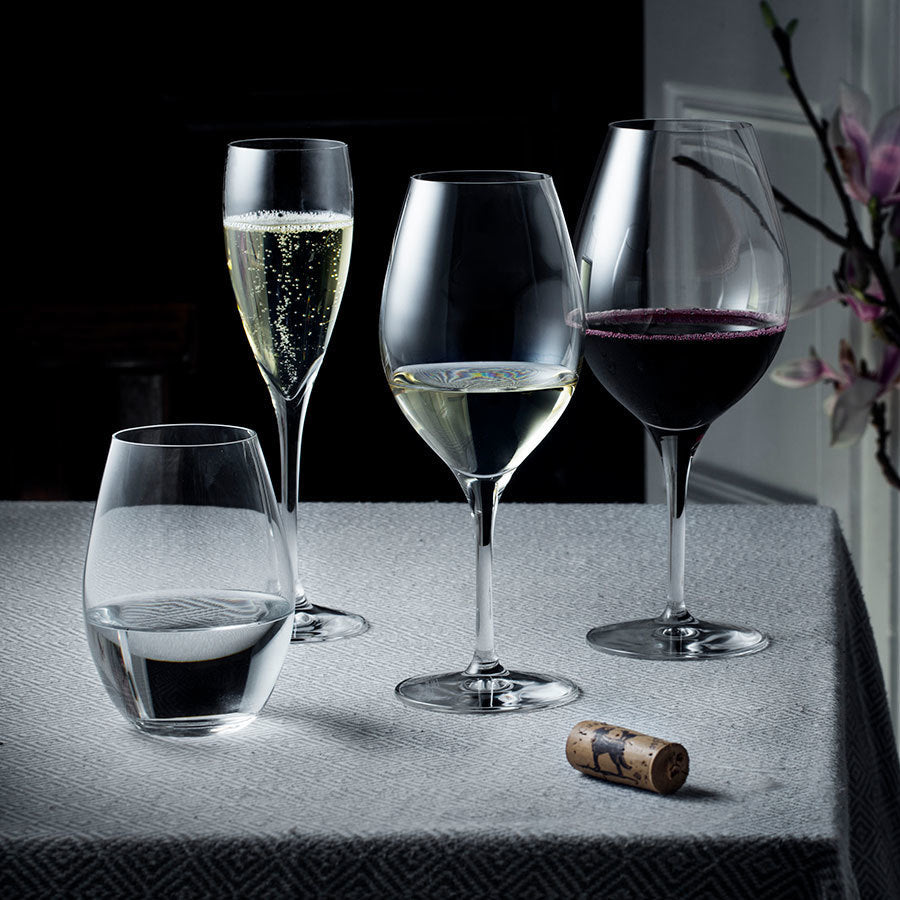 A table features four Orrefors wine glasses, including a Champagne glass from the More series (5oz, 2-pack), possibly designed by Erika Lagerbielke. Each glass contains different beverages with a cork nearby, set against a dark background.