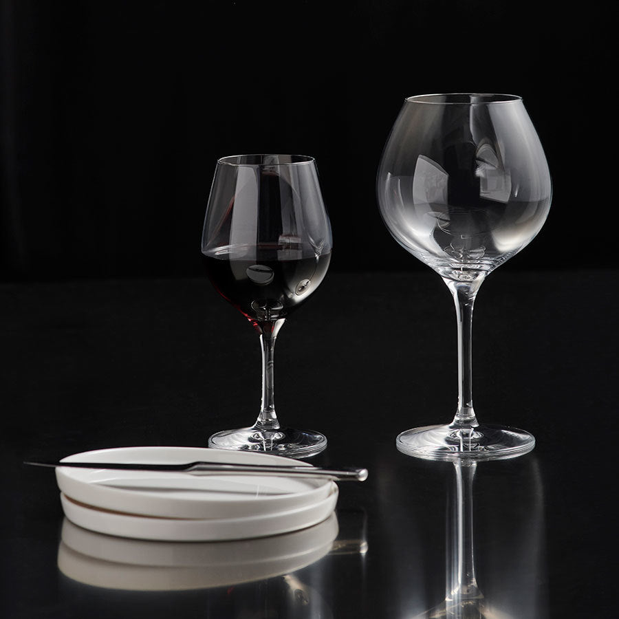 Two empty glasses from the Orrefors: More Bistro Wine Glass (4-Pack) sit beside a stack of white plates with a knife on top, set against a black background.