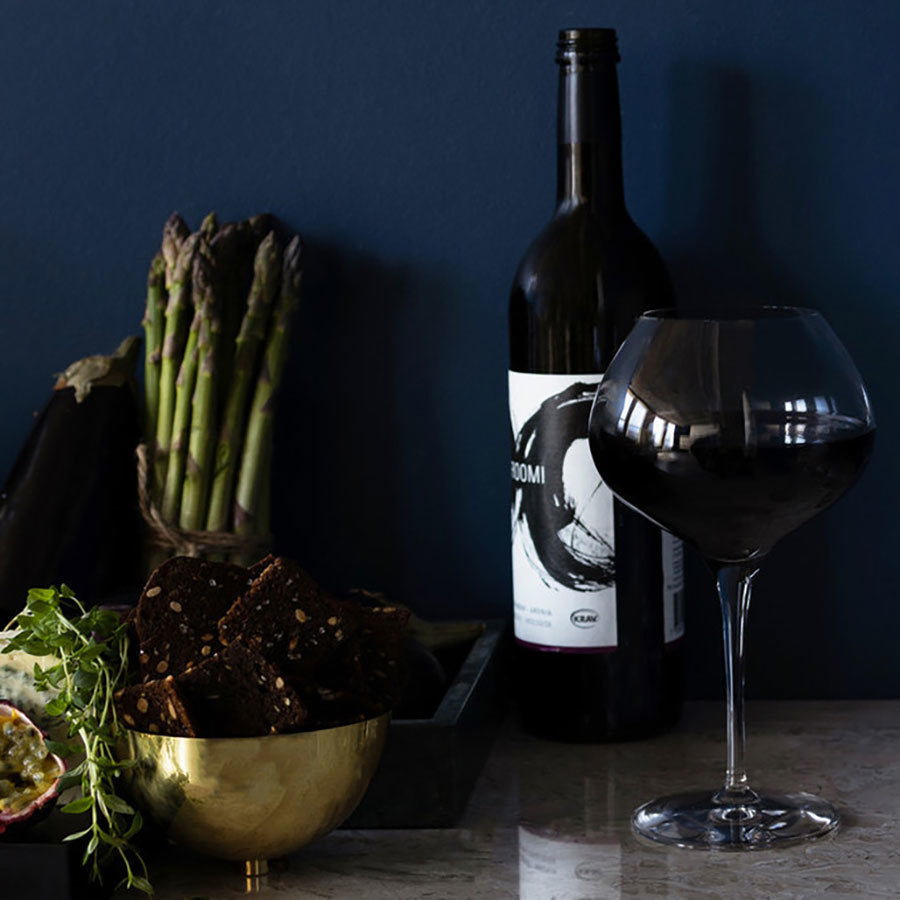 A bottle of red wine and an Orrefors More Mature Glass 16oz rest elegantly on a table. Nearby, asparagus spears, crispy brown crackers in a bowl, a half-cut passion fruit, and fresh herb sprigs complete the setting.