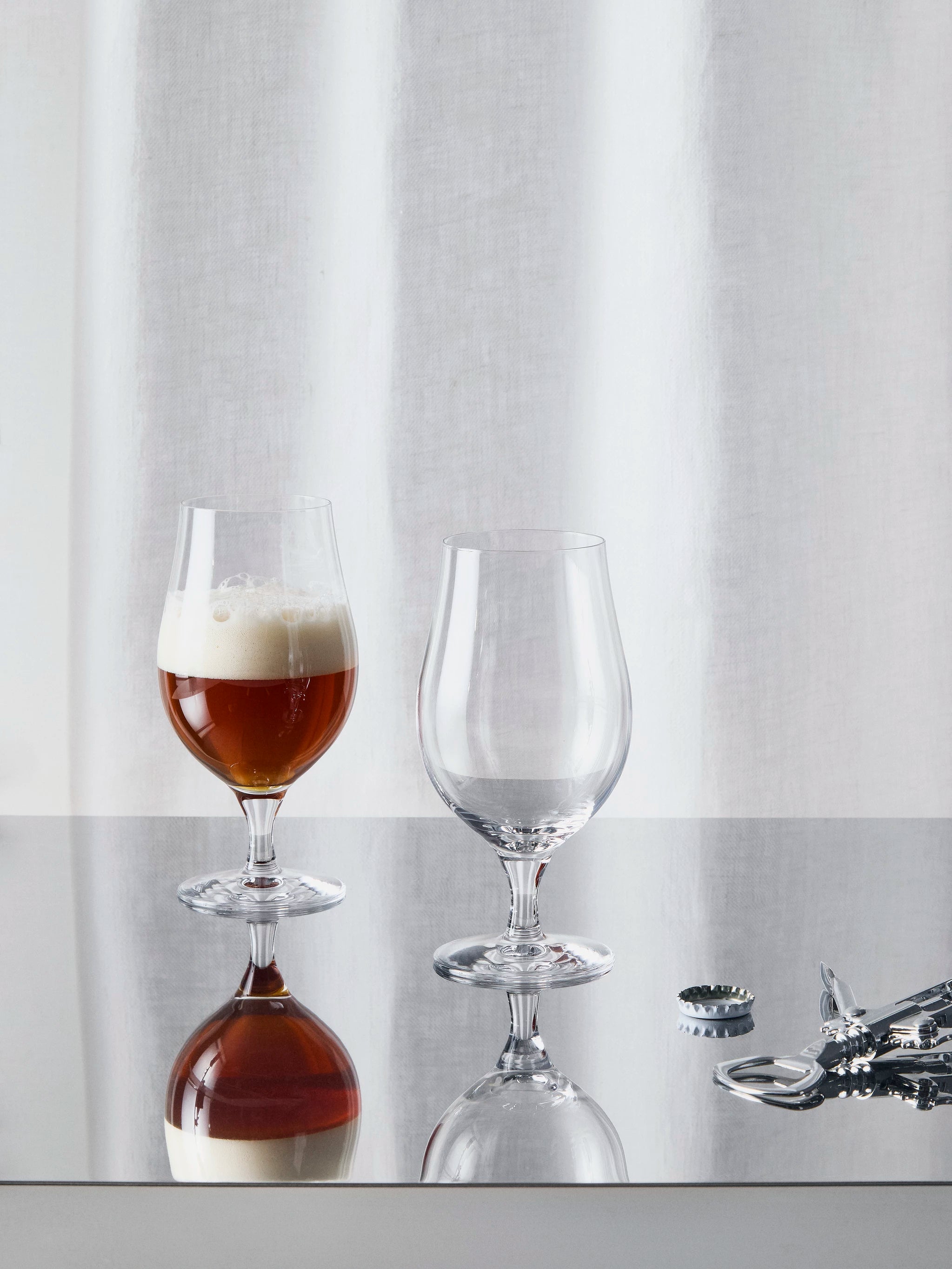 Two Orrefors: More Beer Taster Glasses, each 16oz, sit on a reflective surface—one filled with beer and foam while the other remains pristine. Nearby, bottle caps and an opener await their next use.