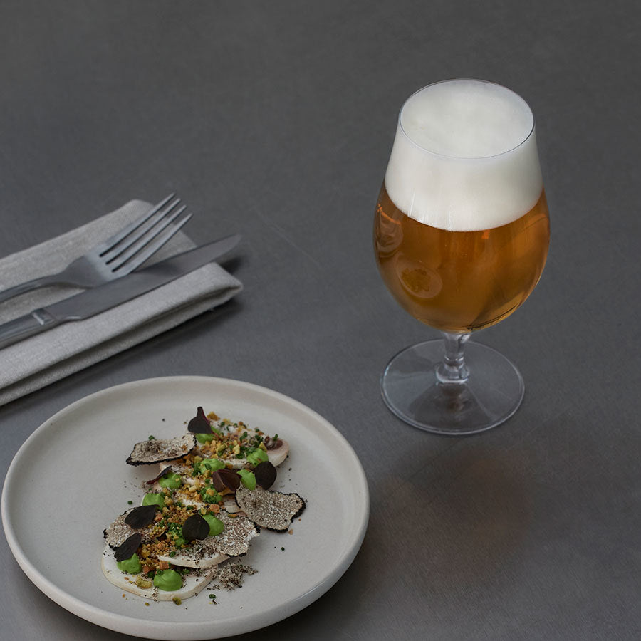 A gourmet dish with truffles and greens is served on a metal table, accompanied by an Orrefors More Beer Taster Glass 16oz (2-Pack). Cutlery rests on a folded napkin beside the plate.