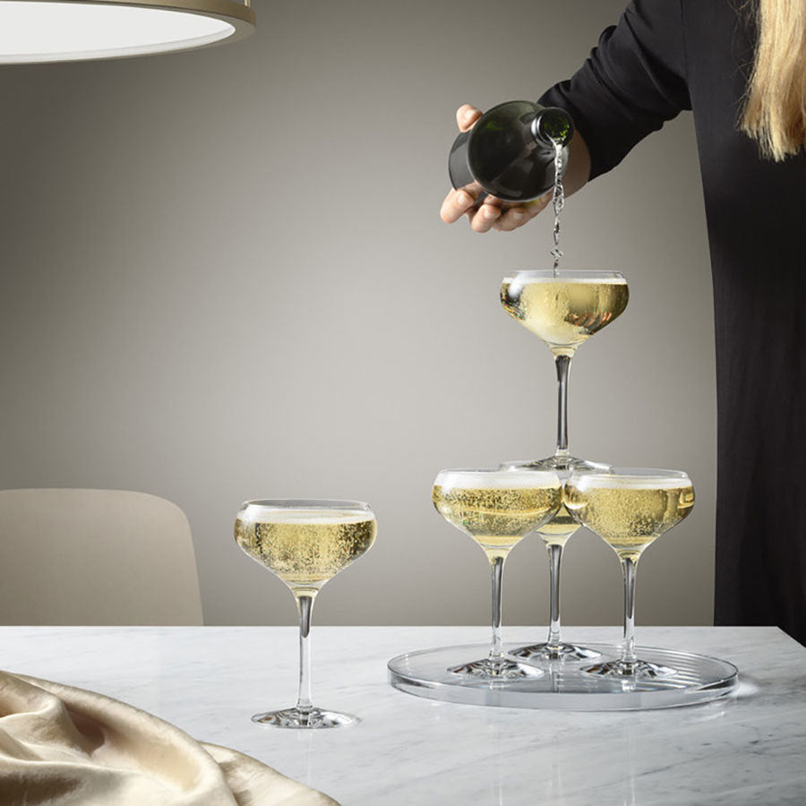 Someone pours champagne into an Orrefors: More Coupe Glass 7oz tower, beautifully set on a white marble table.