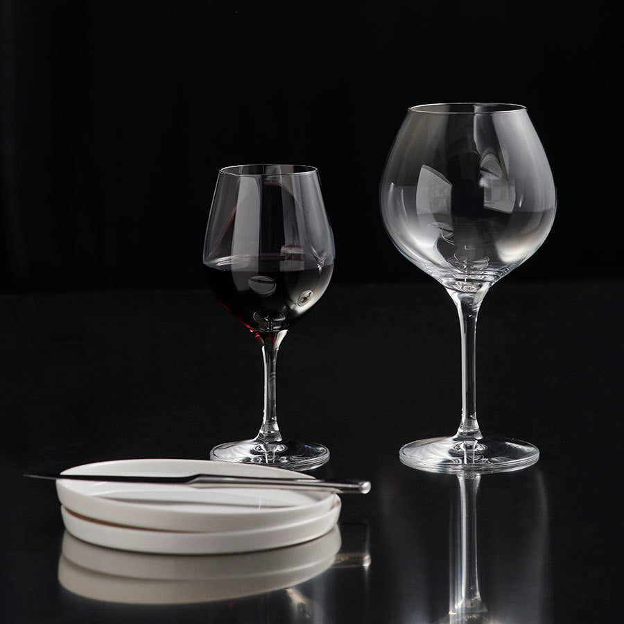 Two Orrefors: More Pinot Glasses, one with red liquid and one empty, sit beside two stacked white plates with a knife on a reflective black surface. Designed by Erika Lagerbielke, these glasses add elegance to any setting.