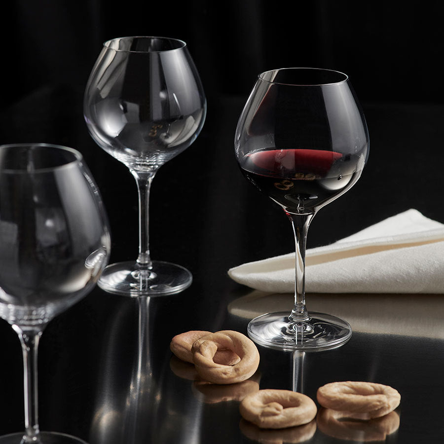 Three Orrefors More Pinot glasses, one with red wine, rest elegantly on a reflective surface next to a folded white napkin and a few round crackers.