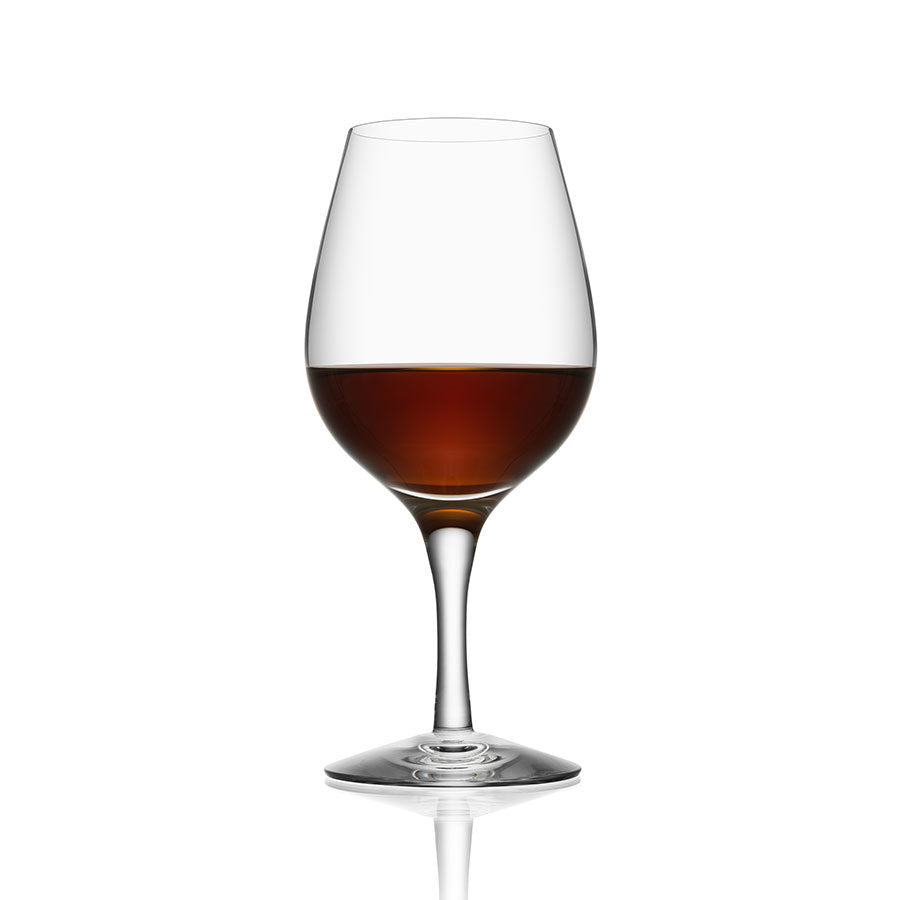 An Orrefors More Spirits glass from the 7oz 4-pack, tulip-shaped and crystal-clear, cradles a rich brown liquid on a reflective white surface.