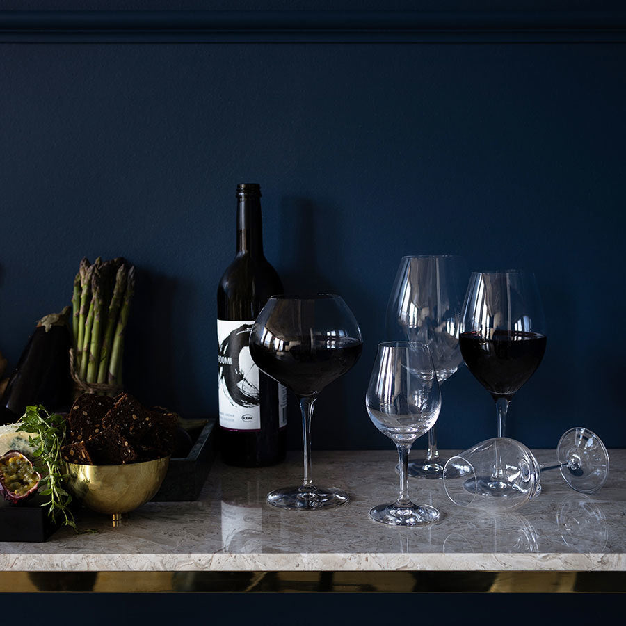 A table set with Orrefors: More Spirits Glass 7oz (4-Pack), a wine bottle, asparagus, and bread rests elegantly on a marble surface against a dark blue wall.