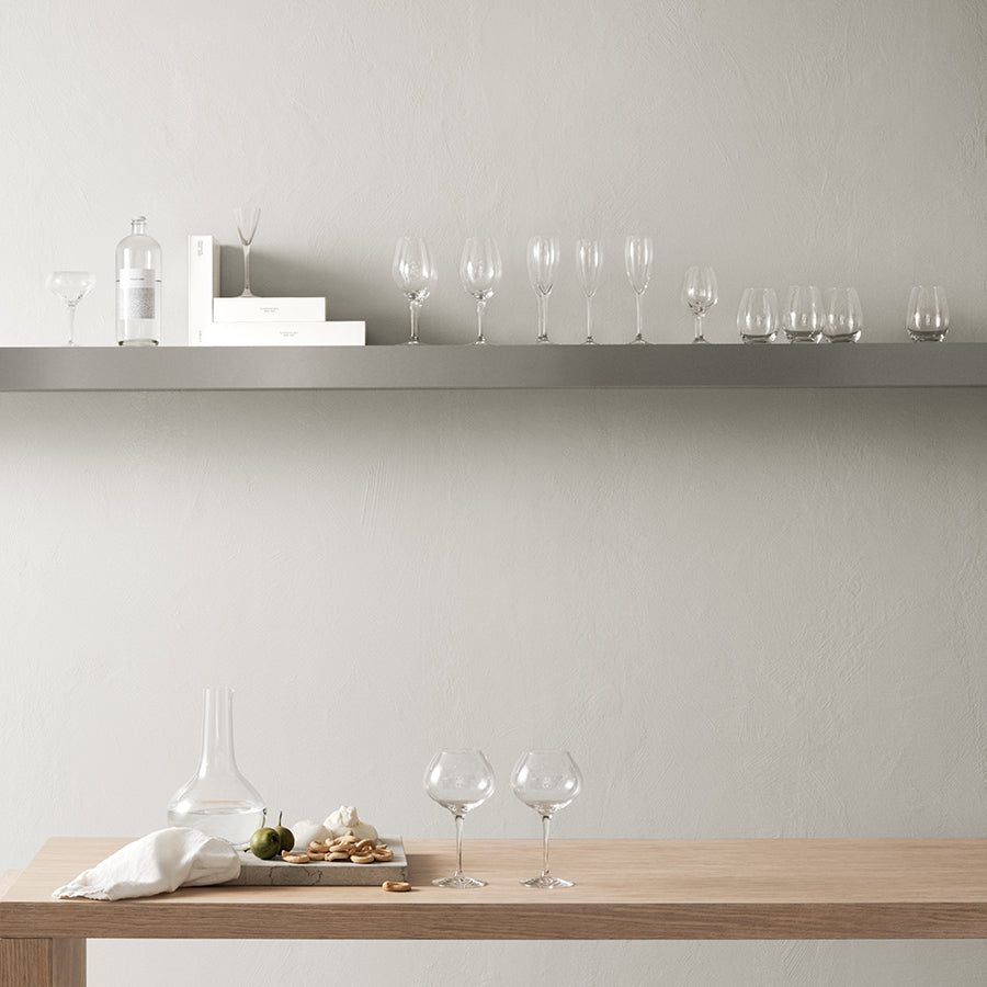 A minimalist interior featuring a wooden table with assorted wine glasses, an Orrefors: More Tumbler 13oz (2-Pack) next to a carafe and cutting board. A floating shelf against a plain wall holds books, a bottle, crystal glassware, and more.