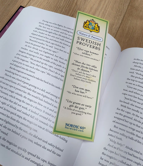 The Bookmark: Swedish Proverbs “Nature & Patience” (2.5" × 8.5") lies on the open pages of a book atop a wooden surface, highlighting its green nature-inspired design and wise Swedish sayings.