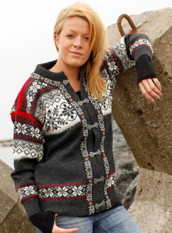 A blonde person in a Norwool Classic Norwegian Cardigan—dark gray, 100% pure wool with Selbu Star design—stands outdoors, leaning against large concrete blocks by a rocky shore.