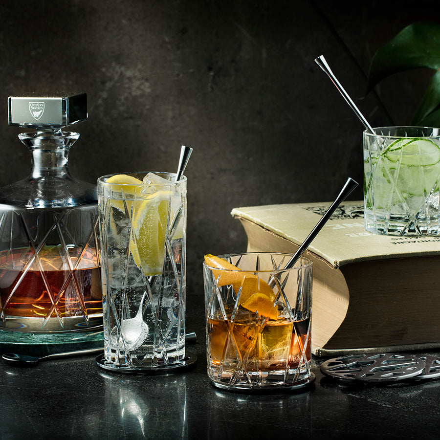 Assorted beverages in Orrefors crystal glasses with metal straws adorn a table beside an Orrefors City Old Fashioned Glass, a decanter, a book, and a decorative plant.