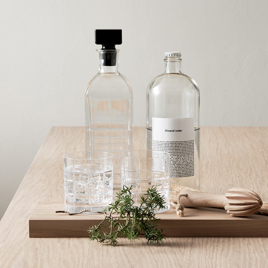 A glass carafe and bottle of mineral water on wood, alongside a 2-pack of Orrefors: Street Double Old Fashioned glasses, a citrus reamer, and a small leafy branch.