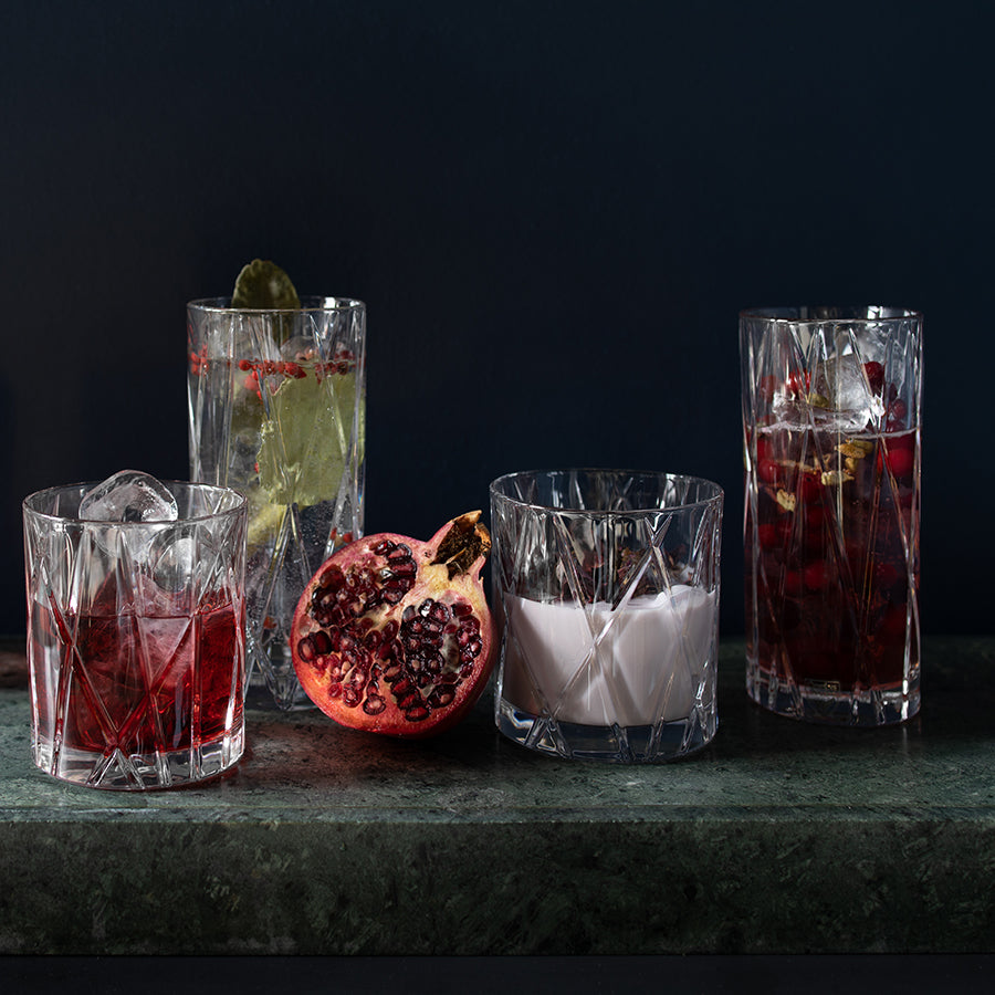 A variety of drinks in Orrefors: City Old Fashioned Glass 8oz (4-Pack) glasses, garnished with ice and lime, surround a halved pomegranate on a dark surface.