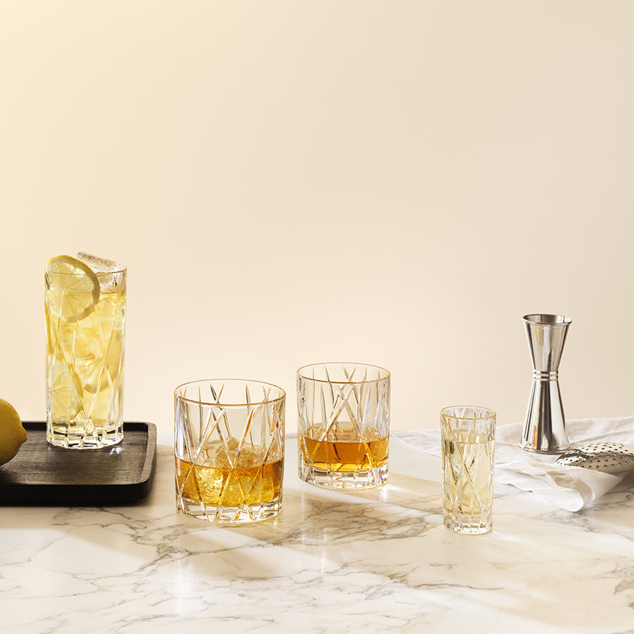 An elegant glassware set with beverages on a marble surface includes highball, Crystal Glass tumblers, and shot glasses. Nearby rests an Orrefors City Old Fashioned Glass 8oz (4-Pack), along with a metal jigger and strainer.