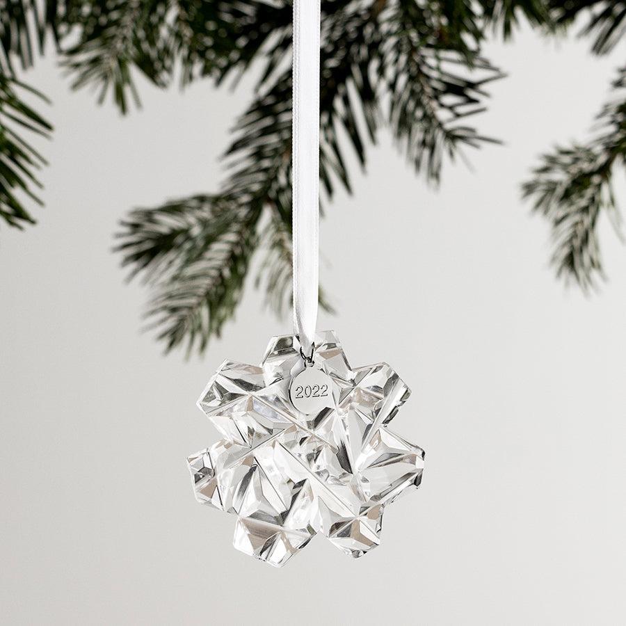 The Orrefors: Annual Ornament Carat, a breathtaking cut crystal snowflake labeled 2022, gracefully hangs from a white ribbon amid lush evergreen branches.