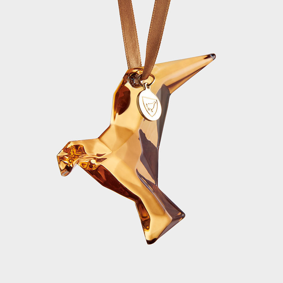 The Orrefors Annual Holiday Ornament, Homage Dove 21K Gold 2024, is a reflective golden origami-style bird pendant with a ribbon loop and a small round tag.