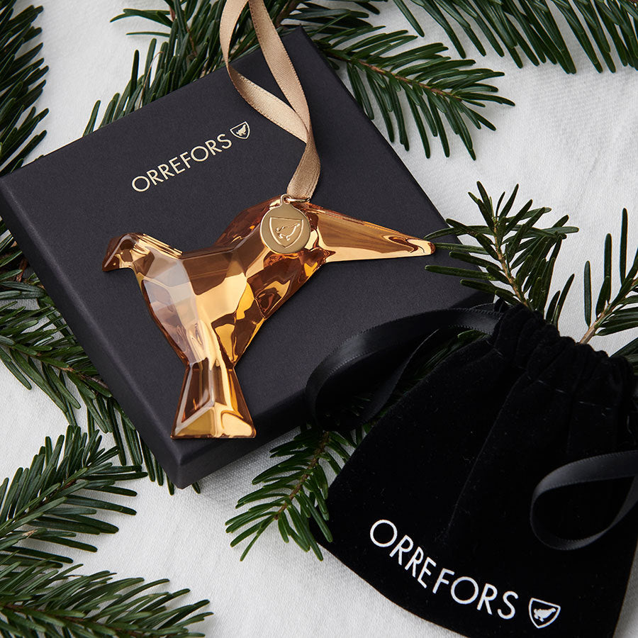 The Orrefors Annual Holiday Ornament, a 21K gold-colored glass dove for 2024, rests elegantly on an Orrefors box with nearby pine branches.