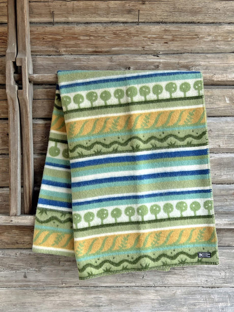 A Kerstin Landstrom Osterlen blanket, made from 100% Norwegian lambs wool and featuring geometric and tree patterns in green, yellow, blue, and white, rests on a wooden surface.