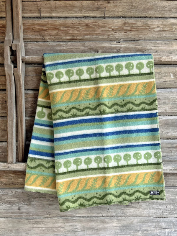 A Kerstin Landstrom Osterlen blanket, made from 100% Norwegian lambs wool and featuring geometric and tree patterns in green, yellow, blue, and white, rests on a wooden surface.