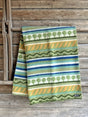 A Kerstin Landstrom Osterlen blanket, made from 100% Norwegian lambs wool and featuring geometric and tree patterns in green, yellow, blue, and white, rests on a wooden surface.