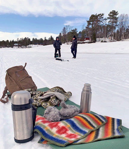 Two people stand on a snowy landscape with trees in the background. In the foreground, a colorful blanket featuring the Rättvik pattern is spread across the snow, accompanied by thermoses, a backpack, scarves, gloves, and an outdoor sports seat pad made of 100% Norwegian Organic Lambswool by Kerstin Landstrom.