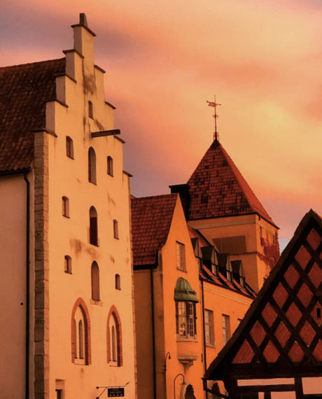 The Visby blanket in rust-colored wool by Kerstin Landström Design captures the essence of Swedish heritage, reminiscent of historic European buildings with steep roofs set against a warm, orange sunset sky, blending seamlessly with the scene's timeless charm.