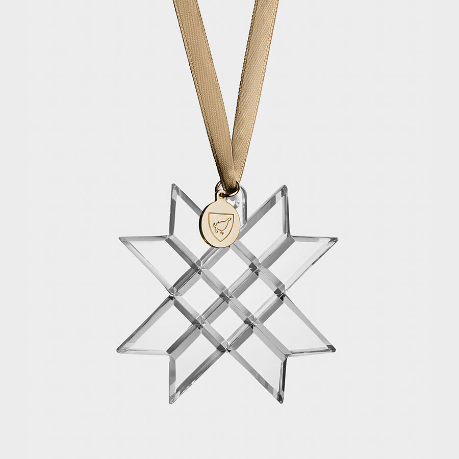 The Orrefors 2023 Annual Ornament features a clear star-shaped crystal with intersecting lines, suspended by a gold ribbon and adorned with a round, gold charm.