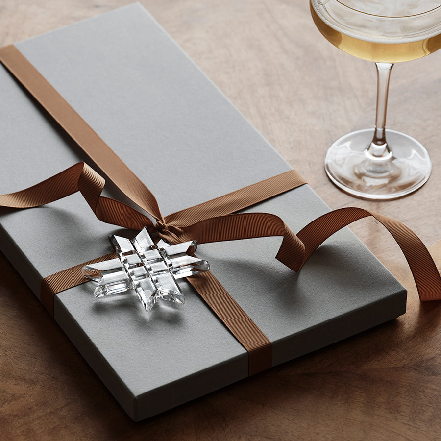 A gray gift box tied with a brown ribbon and an Orrefors: Annual Ornament Paper Star, situated next to a half-filled champagne glass on a wooden surface.