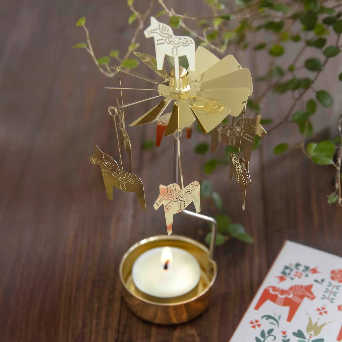Gold-colored Swedish rotary candle holder with hanging Dala horse ornaments above a lit tea light.