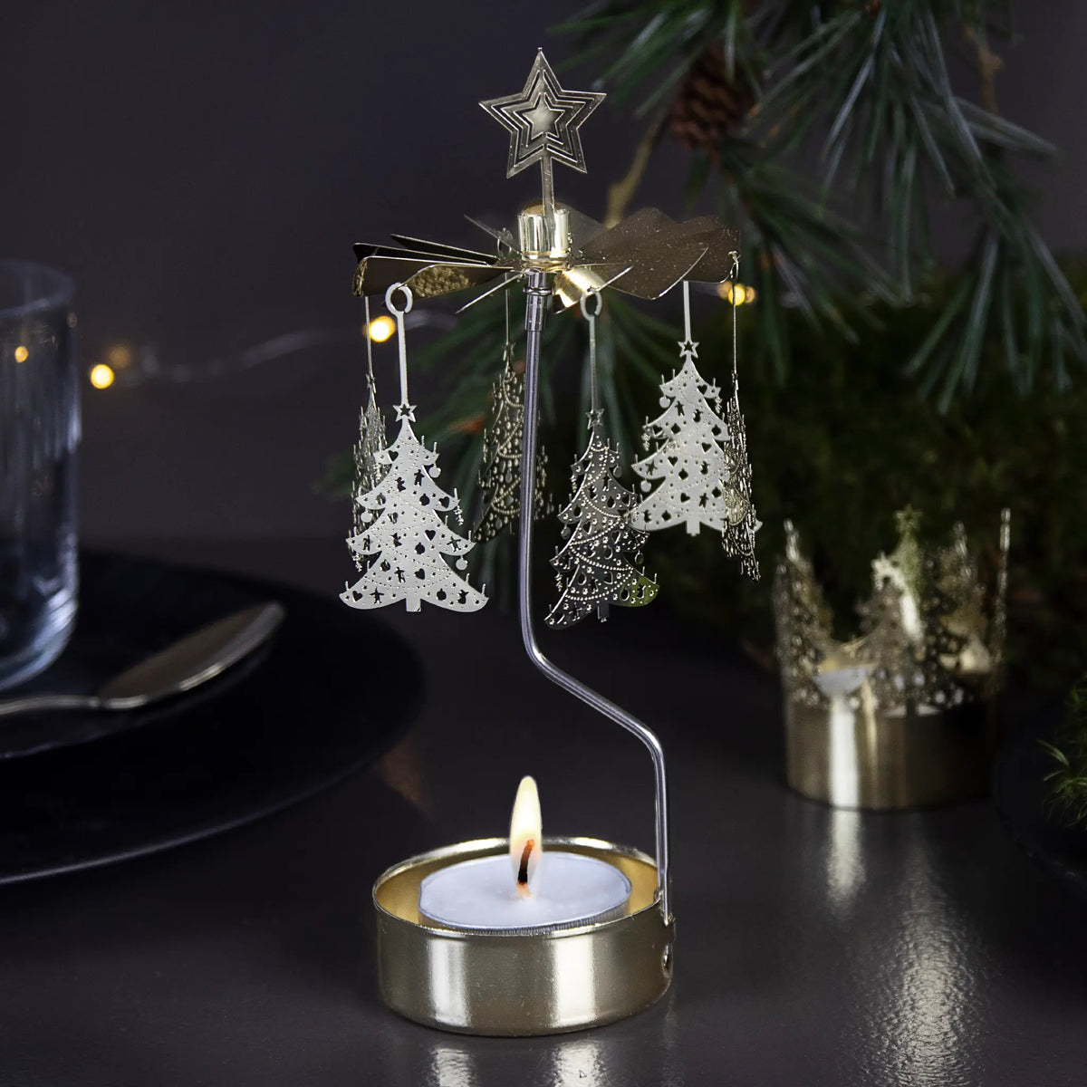 Gold rotary candle holder shaped like a Christmas tree, spinning above a tealight, with hanging ornaments. Festive greenery and string lights blurred in the background.