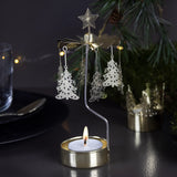 Gold rotary candle holder shaped like a Christmas tree, spinning above a tealight, with hanging ornaments. Festive greenery and string lights blurred in the background.