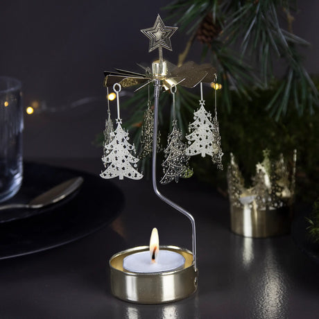 Gold rotary candle holder shaped like a Christmas tree, spinning above a tealight, with hanging ornaments. Festive greenery and string lights blurred in the background.