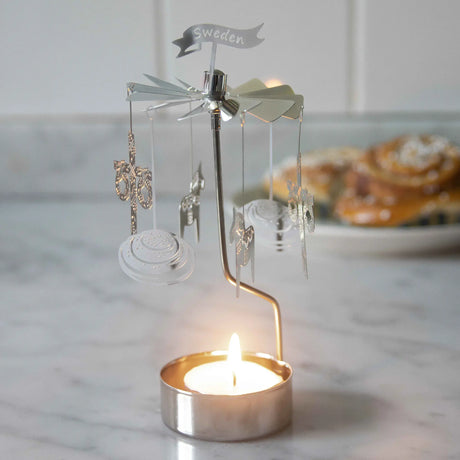 Rotary candle holder with hanging ornaments spinning above a lit tealight on a marble counter, cinnamon buns blurred in the background.
