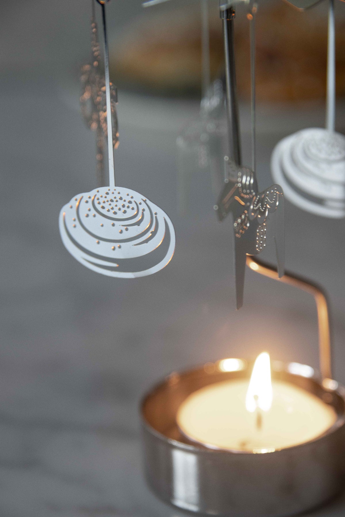Lit tealight candle under a rotary carousel candle holder with white, spiral-shaped ornaments; softly blurred background.