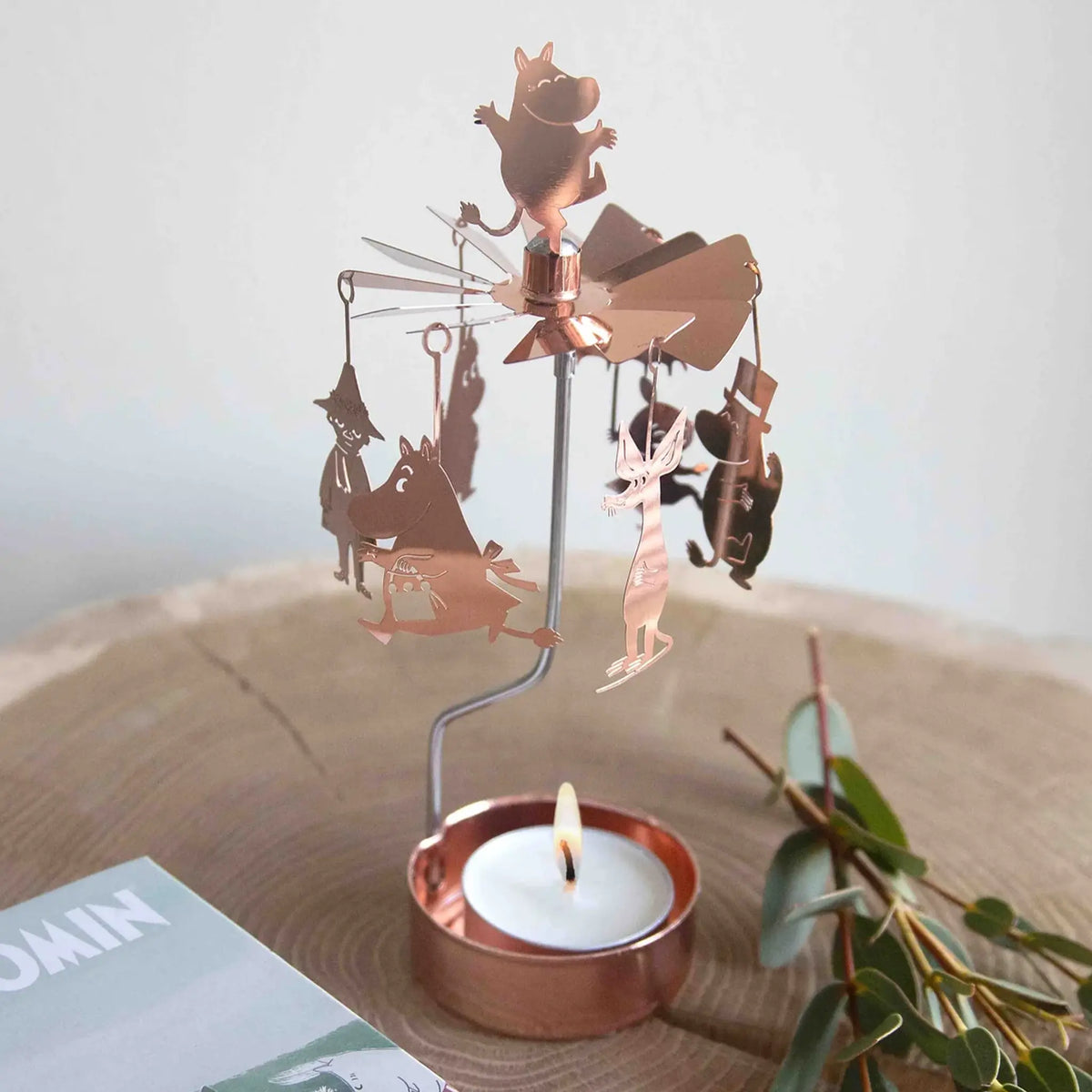 Rose gold copper candle carousel featuring Moomin family figures spinning above a lit tealight, placed on a wooden surface next to greenery and an open book.