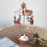 Rose gold copper candle carousel featuring Moomin family figures spinning above a lit tealight, placed on a wooden surface next to greenery and an open book.