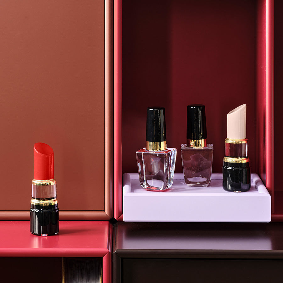 A Kosta Boda Nail Polish in Poppy Red and three nail polish bottles with black caps elegantly rest on a vibrant, multi-colored shelf with pink, brown, and purple sections, reflecting Åsa Jungneliuss chic style.