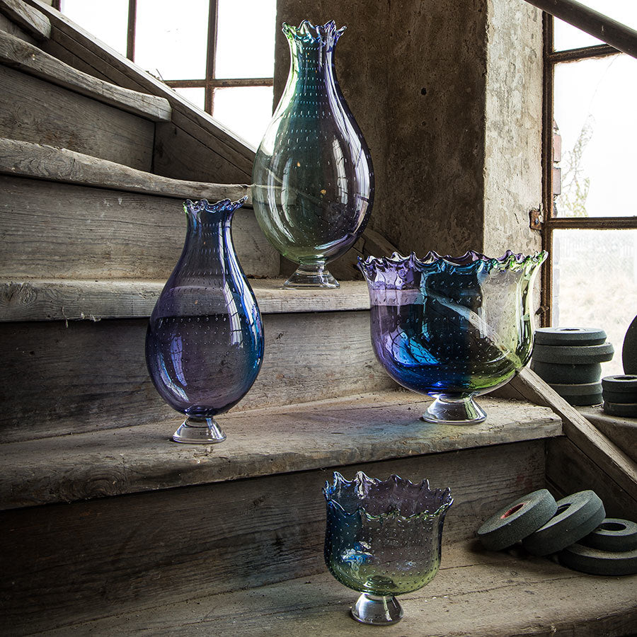 Four multicolored KostaBoda: Poppy Bowl glass vases, evoking Kjell Engmans vibrant designs, adorn the wooden stairs near a rustic wall and windows.