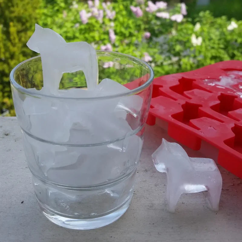 A glass containing horse-shaped ice cubes, one perched on top, features charming Dala Horses created by the red Candy Mold: Dalahast Pralinform, with green foliage enhancing the natural ambiance in the background.