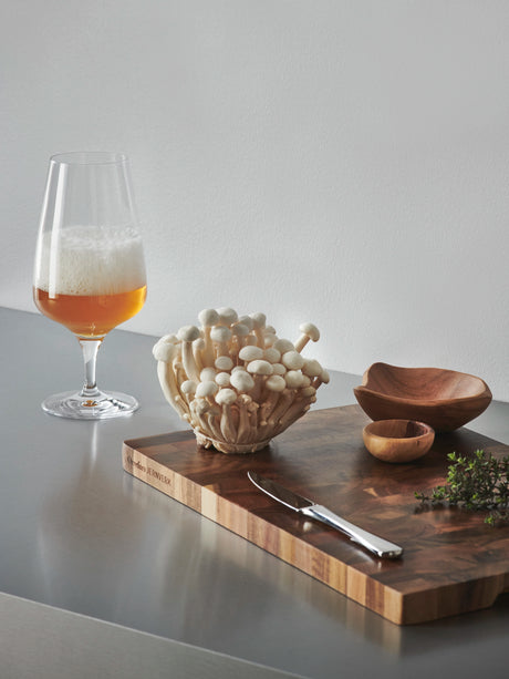 A glass of beer in an Orrefors: Pulse Beer Glass 18.5oz (4-Pack) by Ingegerd Raman sits alongside white mushrooms, a knife, wooden bowls, and fresh herbs, showcasing Scandinavian simplicity on the counter.