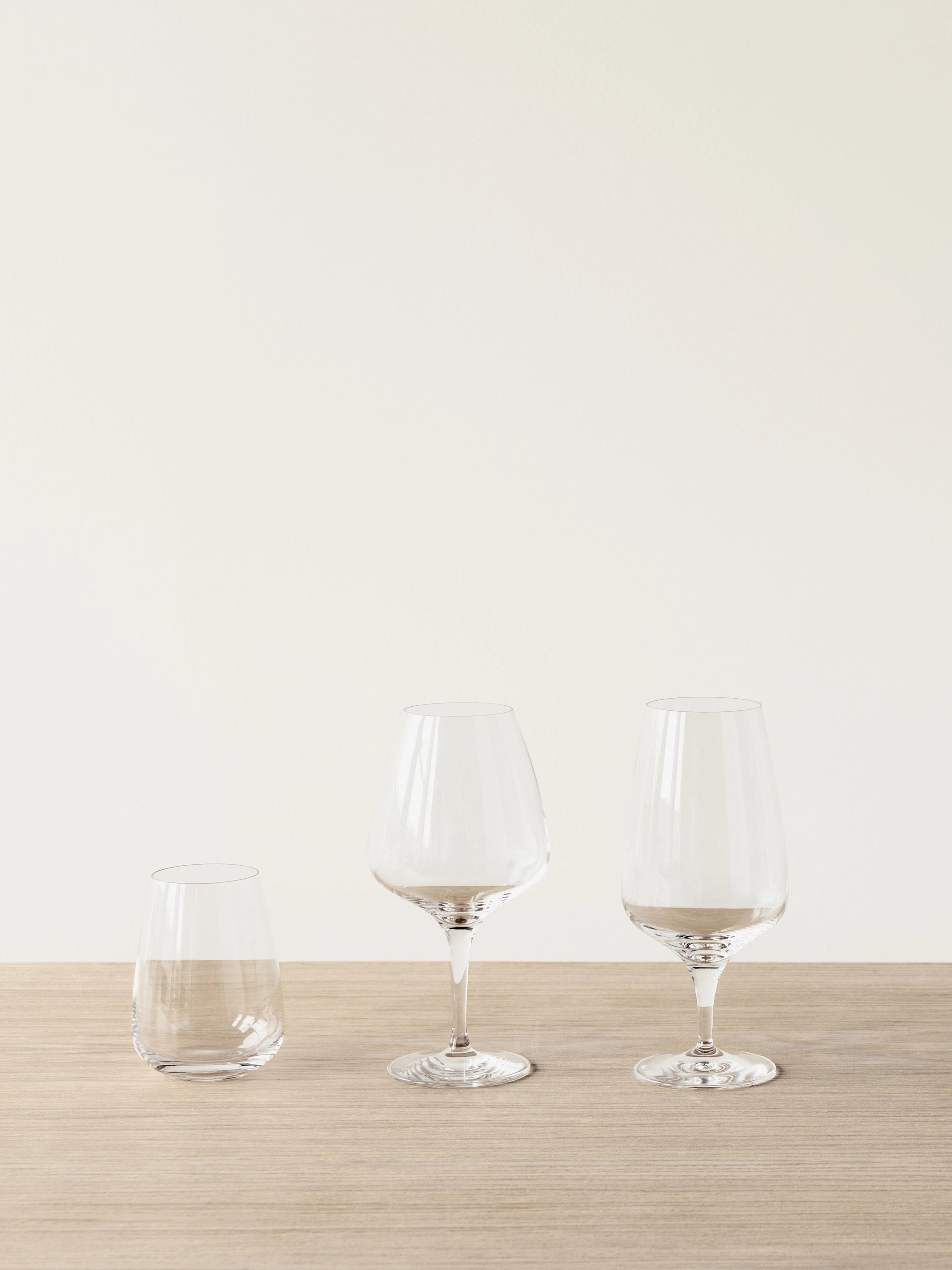 Three crystal glasses are placed on a wooden surface against a plain backdrop, featuring an Orrefors Pulse Beer Glass 18.5oz on the left, flanked by two taller wine glasses to the right.