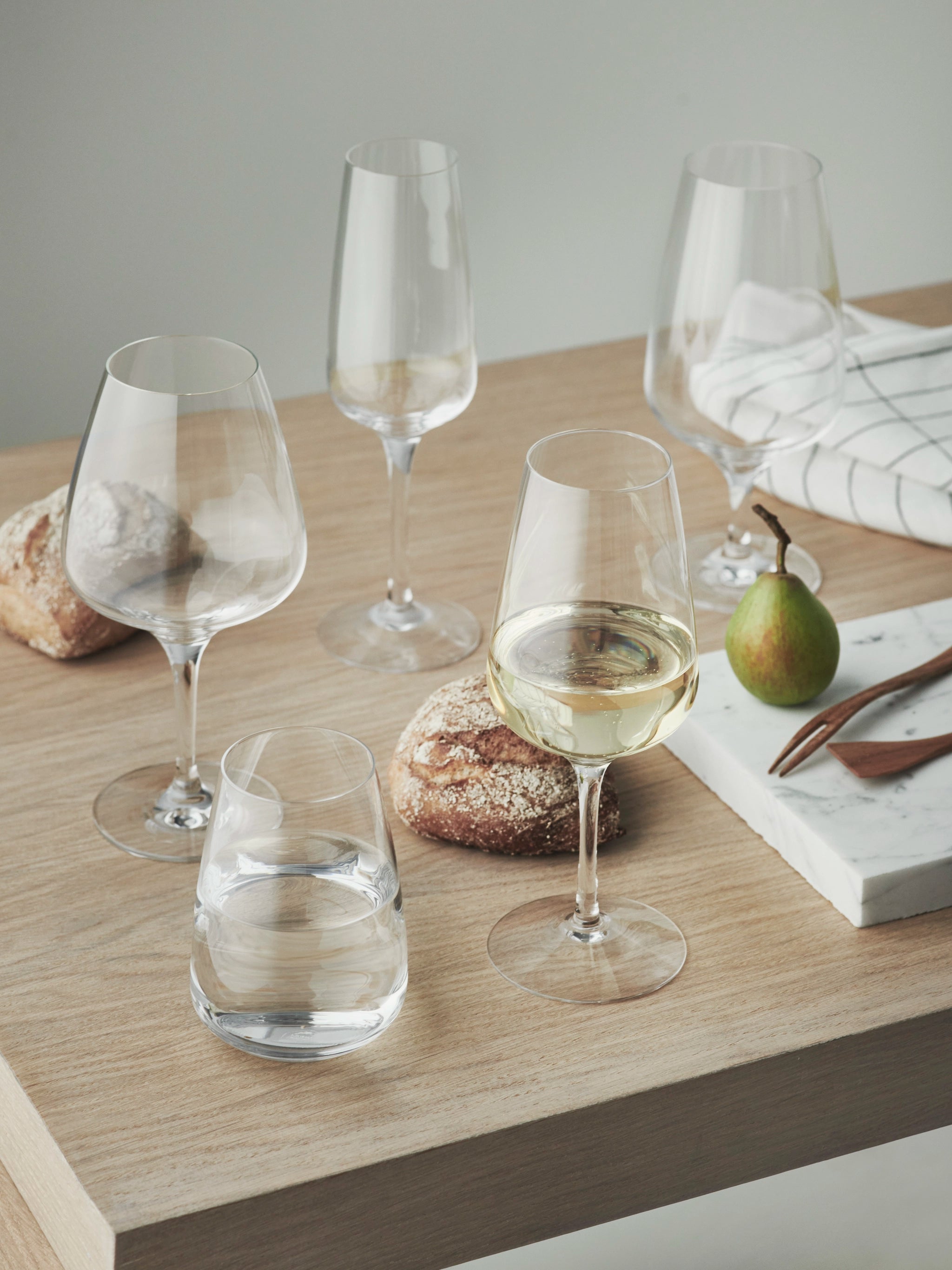 A wooden table is elegantly set with four Orrefors Pulse beer glasses, a glass of white wine, two crusty bread rolls, a pear, and a marble slab adorned with utensils.