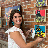 A woman smiles while hanging the vibrant "Nature's Notes 1000 Piece 'Mixed Up!'" PuzzleTwist jigsaw puzzle by Sarah Angst on a brick wall among other colorful animal artworks, featuring the distinctive Maynards style.