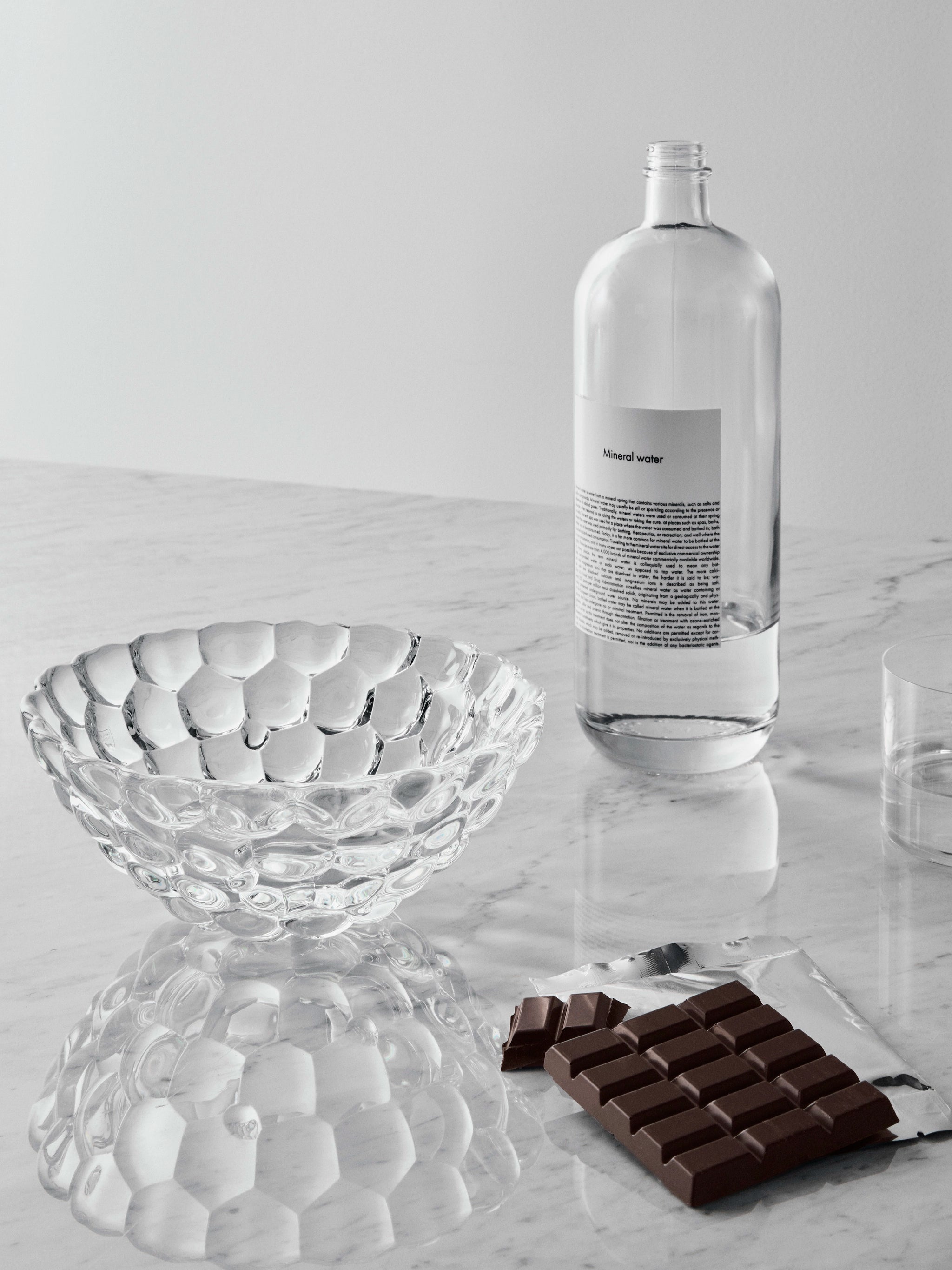 An Orrefors Raspberry Bowl Small, a bottle of mineral water, a chocolate bar, and a crystal glass sit elegantly on the marble surface.