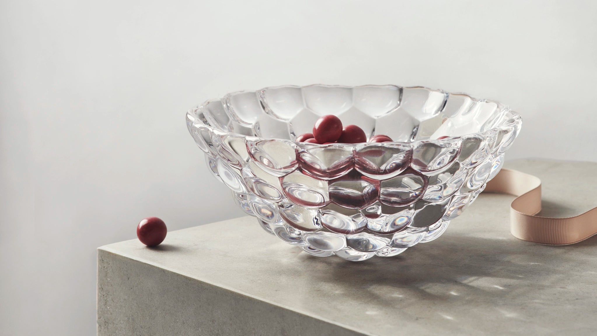 An Orrefors: Raspberry Bowl Small, adorned with a textured glass reminiscent of raspberry patterns, holds red cherries with one outside on the concrete. A light pink ribbon from the right adds softness to the scene.