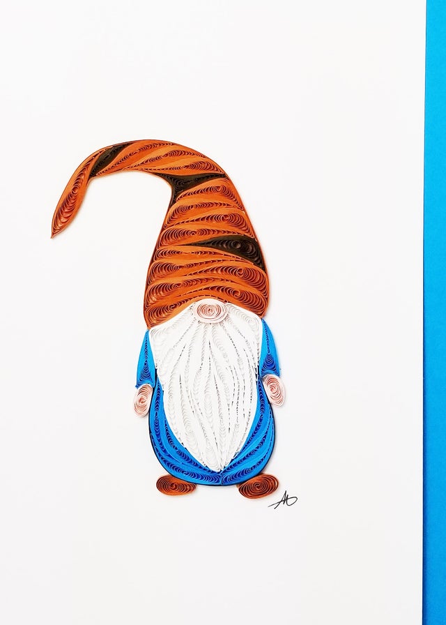 The Cards: Gnome with Brown Hat Quilling Card features exquisite handcrafted quilling with high-quality paper, showcasing a charming gnome adorned in a tall brown hat. Its white beard and blue body stand out beautifully against a white background framed by a partial blue border.