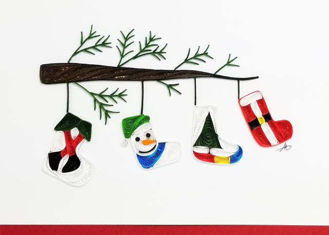 The Card: Holiday Stockings on a Tree Branch Quilling Card 5x7 features intricate paper quilling of a branch with four festive ornaments—a mitten, snowman, stocking, and Santa suit—beautifully capturing holiday cheer.