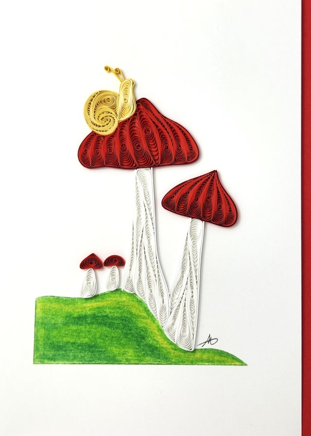 The Card: Mushrooms with Snail Quilling Card 5x7 is a handmade 3D art card featuring quilled red mushrooms with white stems on green grass and a yellow snail, all set against a plain white background.