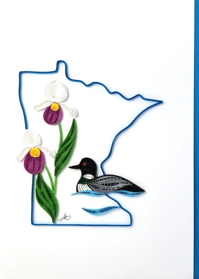 The Minnesota Lady Slipper & Loon Quilling card showcases intricate quilled paper art of the state's bird, the loon, and flower, lady's slipper, elegantly framed by a blue silhouette of Minnesota. It's an ideal card for nature enthusiasts.