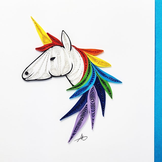 The "Unicorn Quilling Card 4x4" is a meticulously hand-crafted quilling card showcasing a unicorn with a radiant rainbow mane set against a white backdrop, elegantly encased by a blue border.